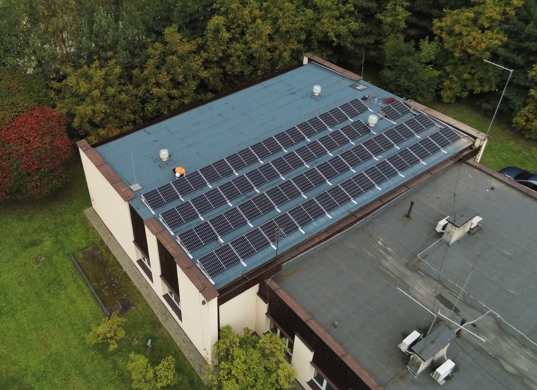 Widok z góry na budynek z zainstalowanymi panelami słonecznymi; pracownik w pomarańczowej kamizelce na dachu podczas inspekcji.