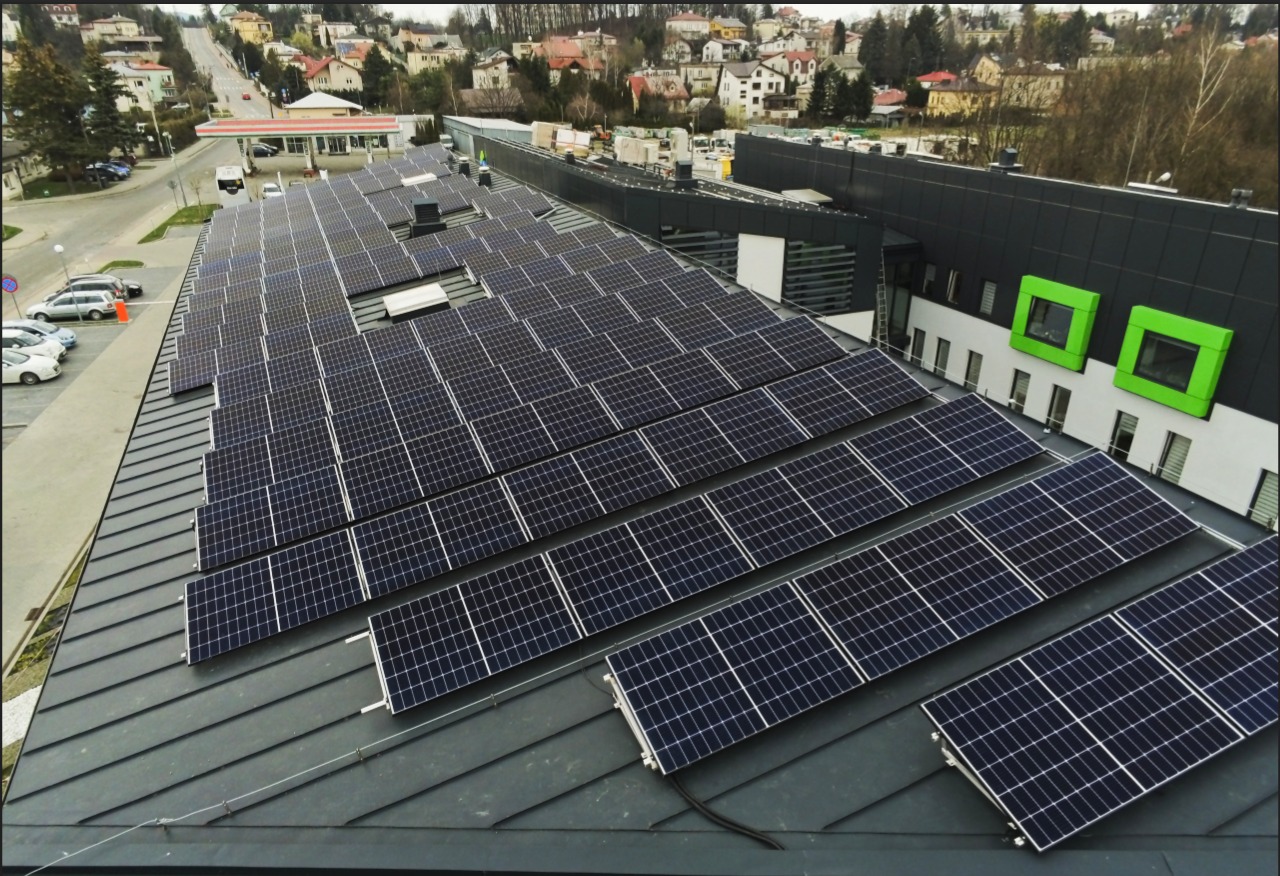 Rozległa instalacja paneli słonecznych na dachu budynku komercyjnego z zielonymi obramowaniami okien, widok z góry na tle zabudowy miejskiej.