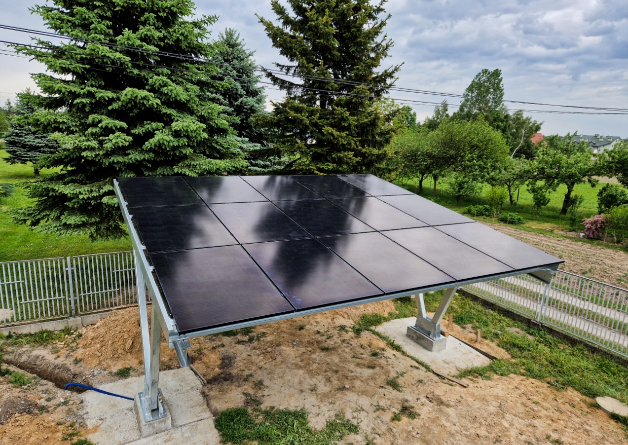 Zainstalowane panele na metalowej konstrukcji w ogrodzie, widoczne drzewa iglaste w tle i częściowo odsłonięta ziemia wokół fundamentów.
