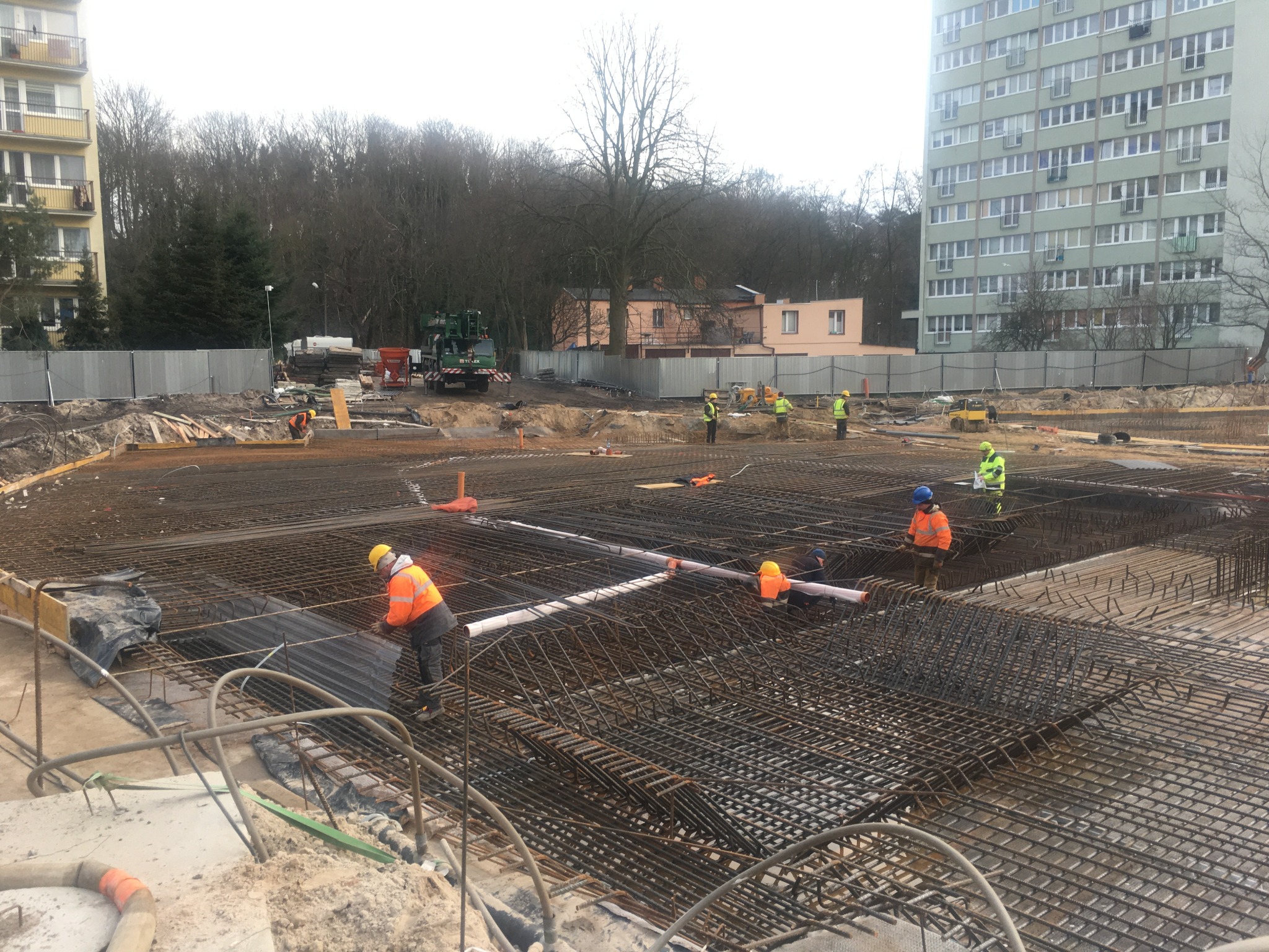Siatka zbrojeniowa na fundamencie budynku wielorodzinnego, robotnicy w odblaskowych ubraniach i kaskach, widok na teren budowy z betonowymi elementami i rurami PCV.