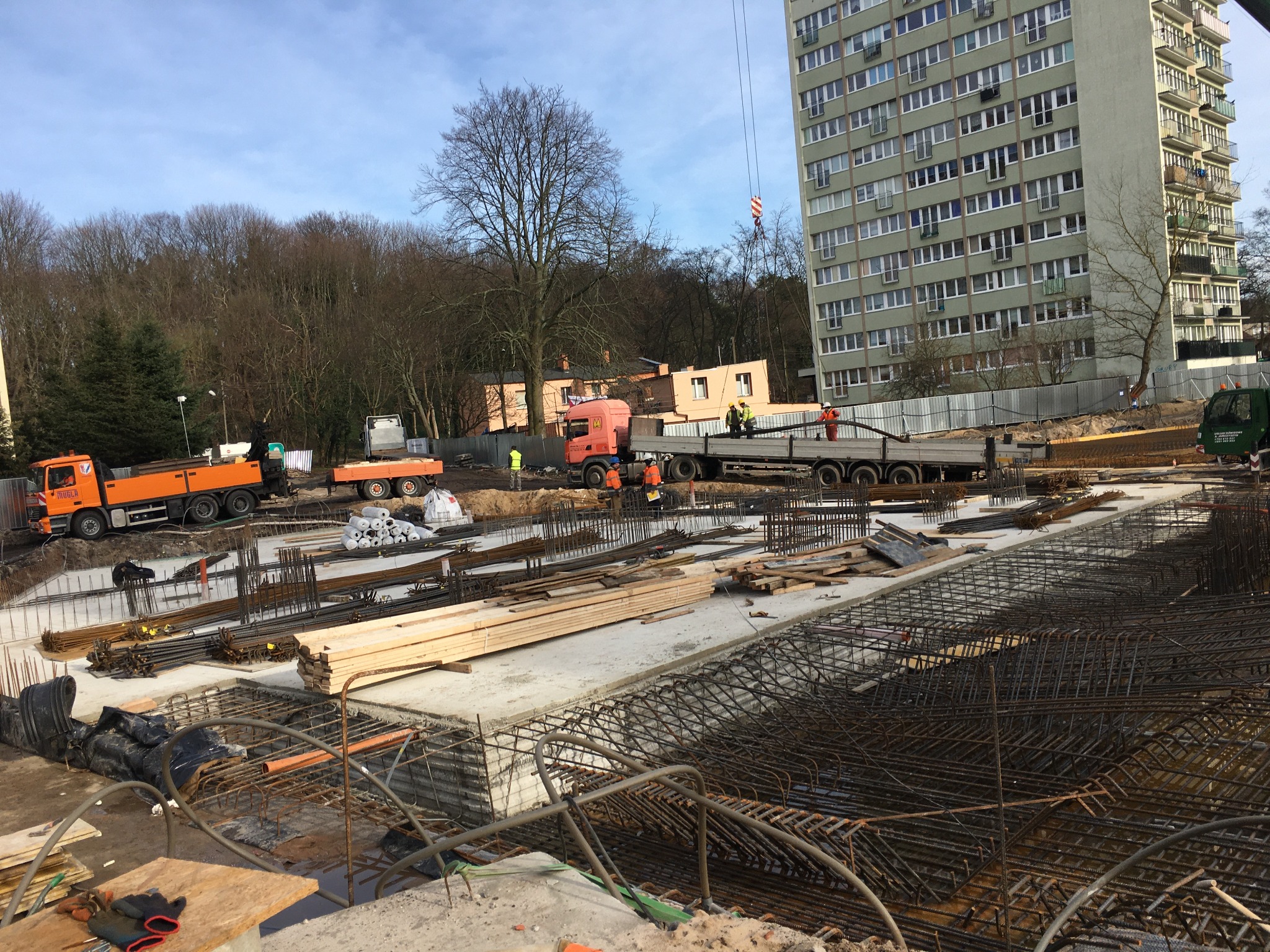 Rozległy plac budowy z widocznym zbrojeniem fundamentów, ciężarówkami i dźwigiem na tle bloku mieszkalnego i drzew