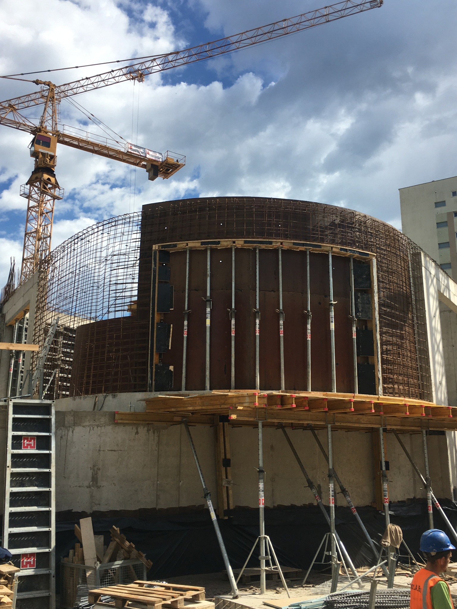 Budowa budynku o zaokrąglonym kształcie z widocznym szkieletem zbrojeniowym, deskowaniem i podporami, z żurawiem wieżowym w tle na tle pochmurnego nieba.