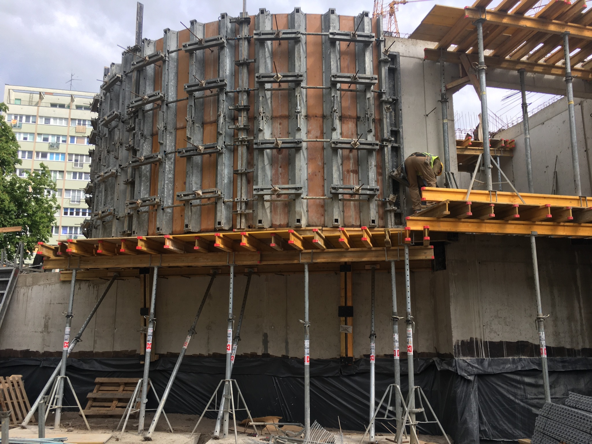 Konstrukcja szalunkowa o okrągłym kształcie na placu budowy, wsparta na podporach, z pracującym robotnikiem w kamizelce odblaskowej, w tle budynek mieszkalny i żuraw budowlany.