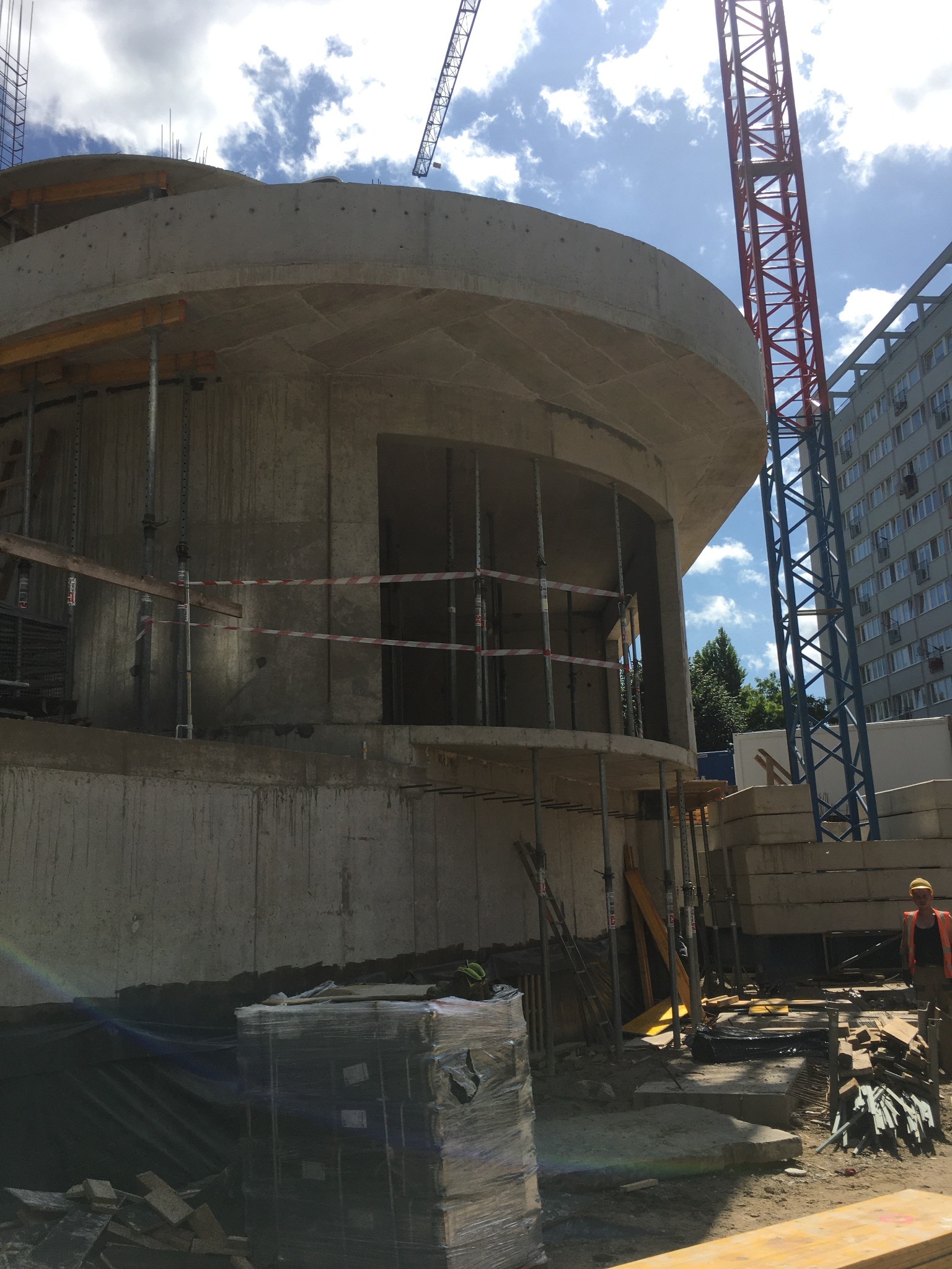 Budowa okrągłego budynku żelbetowego w trakcie realizacji, widoczne szalunki, rusztowania, dźwig i pracownik w kamizelce odblaskowej.