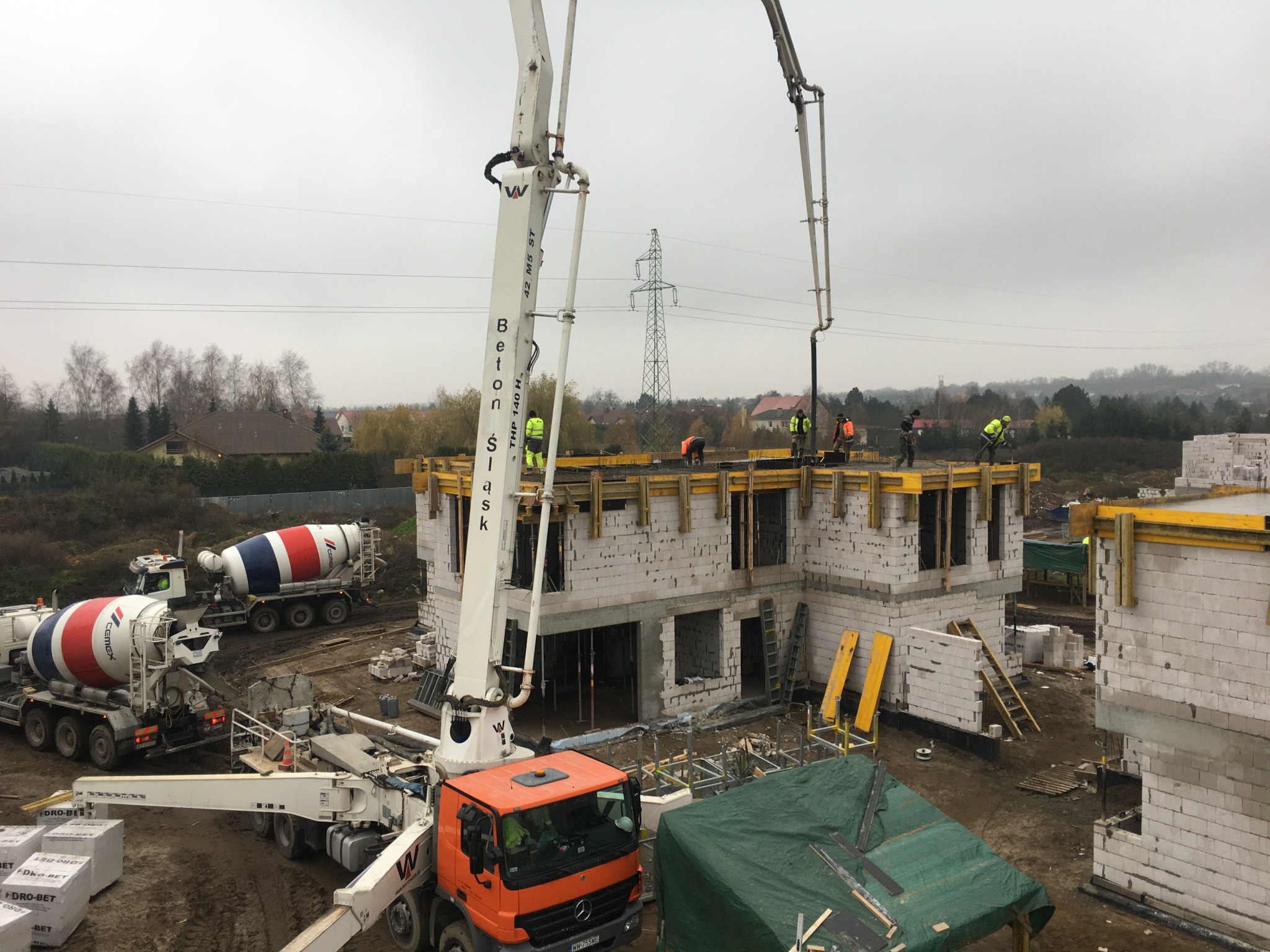 Budowa domu z betonu komórkowego: wylany fundament, ściany parteru i szalunki stropu, pompy do betonu, betoniarki CEMEX, pracownicy w odblaskowych kamizelkach. Widok z góry na plac budowy...