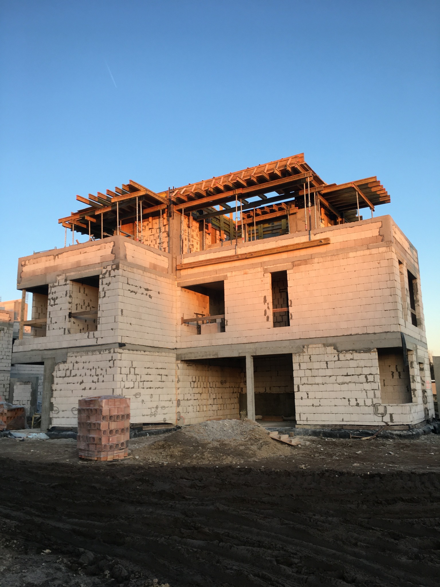 Surowy stan dwukondygnacyjnego budynku z betonu komórkowego w trakcie budowy, z widocznym szalunkiem drewnianym na dachu, sfotografowany przy zachodzącym słońcu.