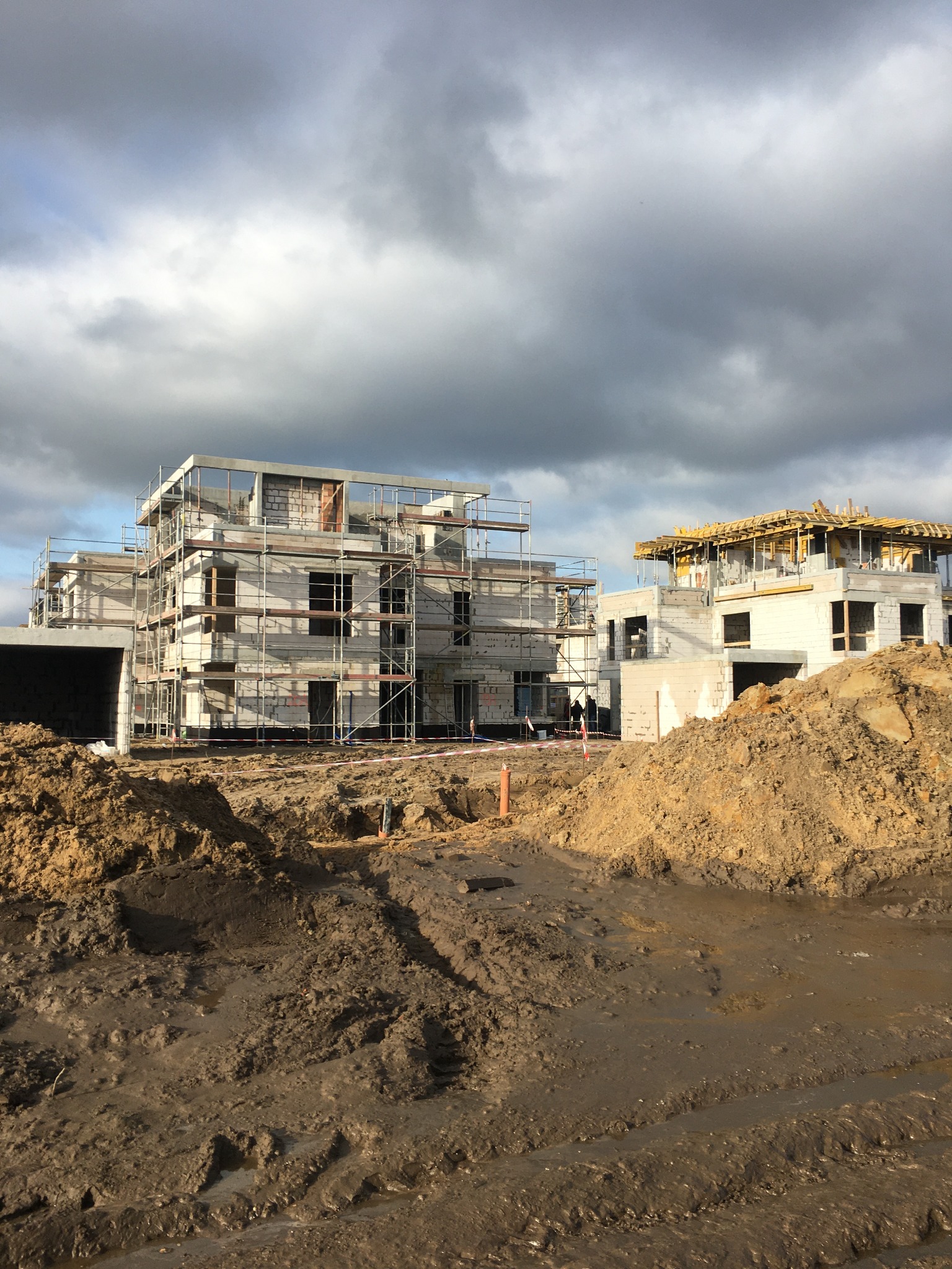 Budowa dwóch budynków w stanie surowym otwartym, z widocznymi rusztowaniami, ziemią i pochmurnym niebem w tle.