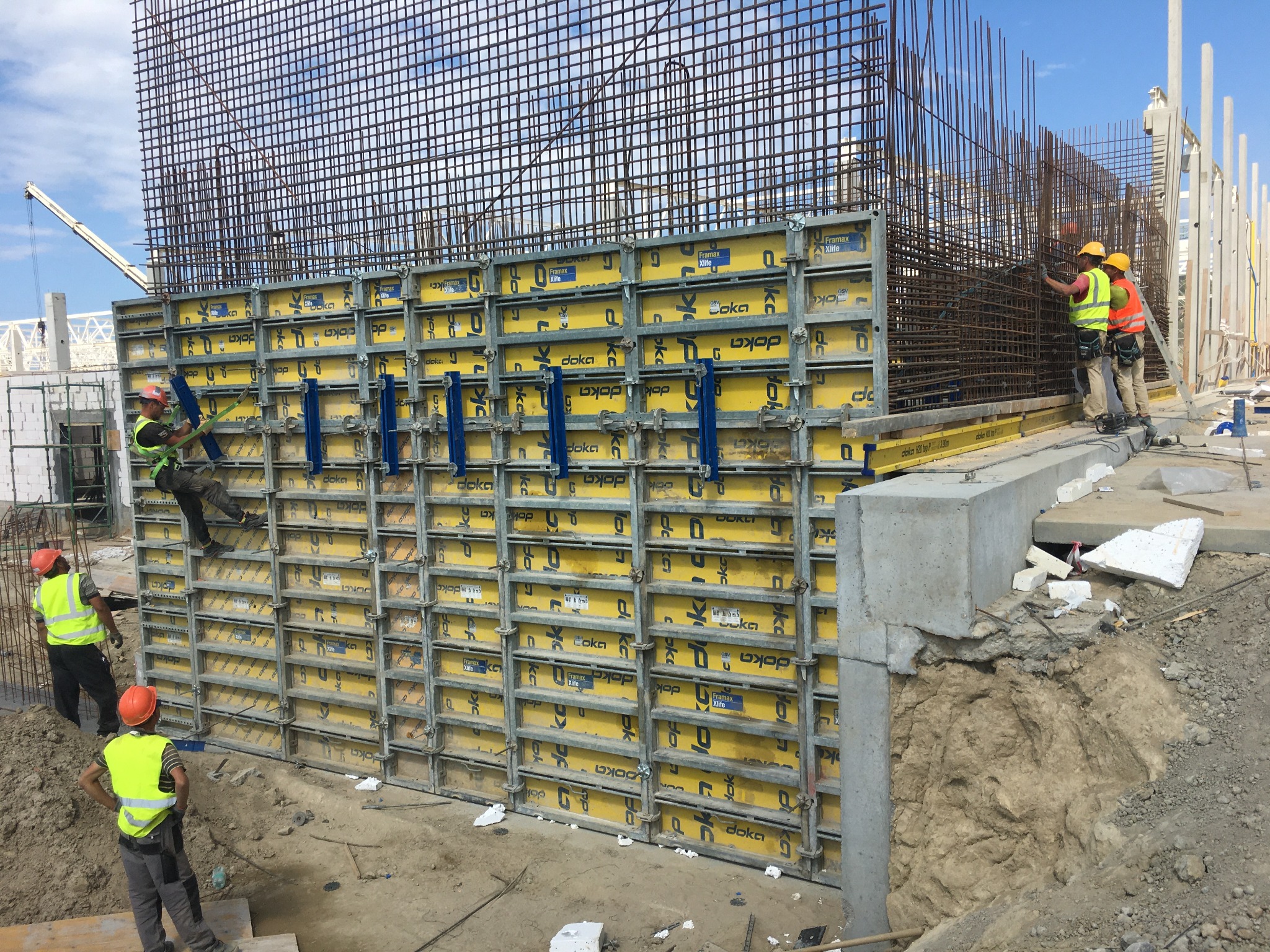 Montaż szalunków Doka i zbrojenia pionowej ściany żelbetowej na placu budowy, widoczni pracownicy w kaskach i kamizelkach odblaskowych, w tle dźwig i konstrukcja stalowa.