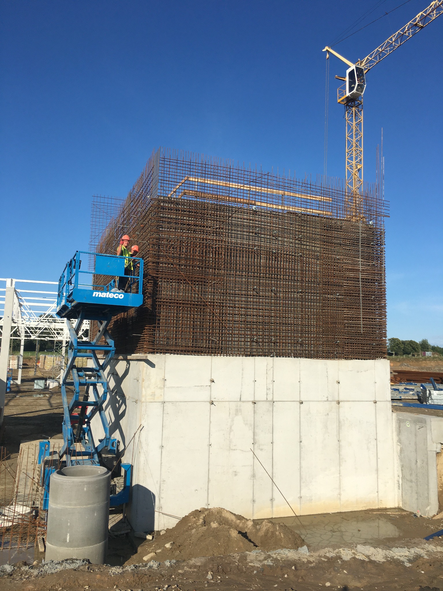 Konstrukcja zbrojeniowa na fundamencie betonowym, widoczni dwaj pracownicy w pomarańczowych kaskach na podnośniku koszowym, w tle żuraw budowlany i błękitne niebo.