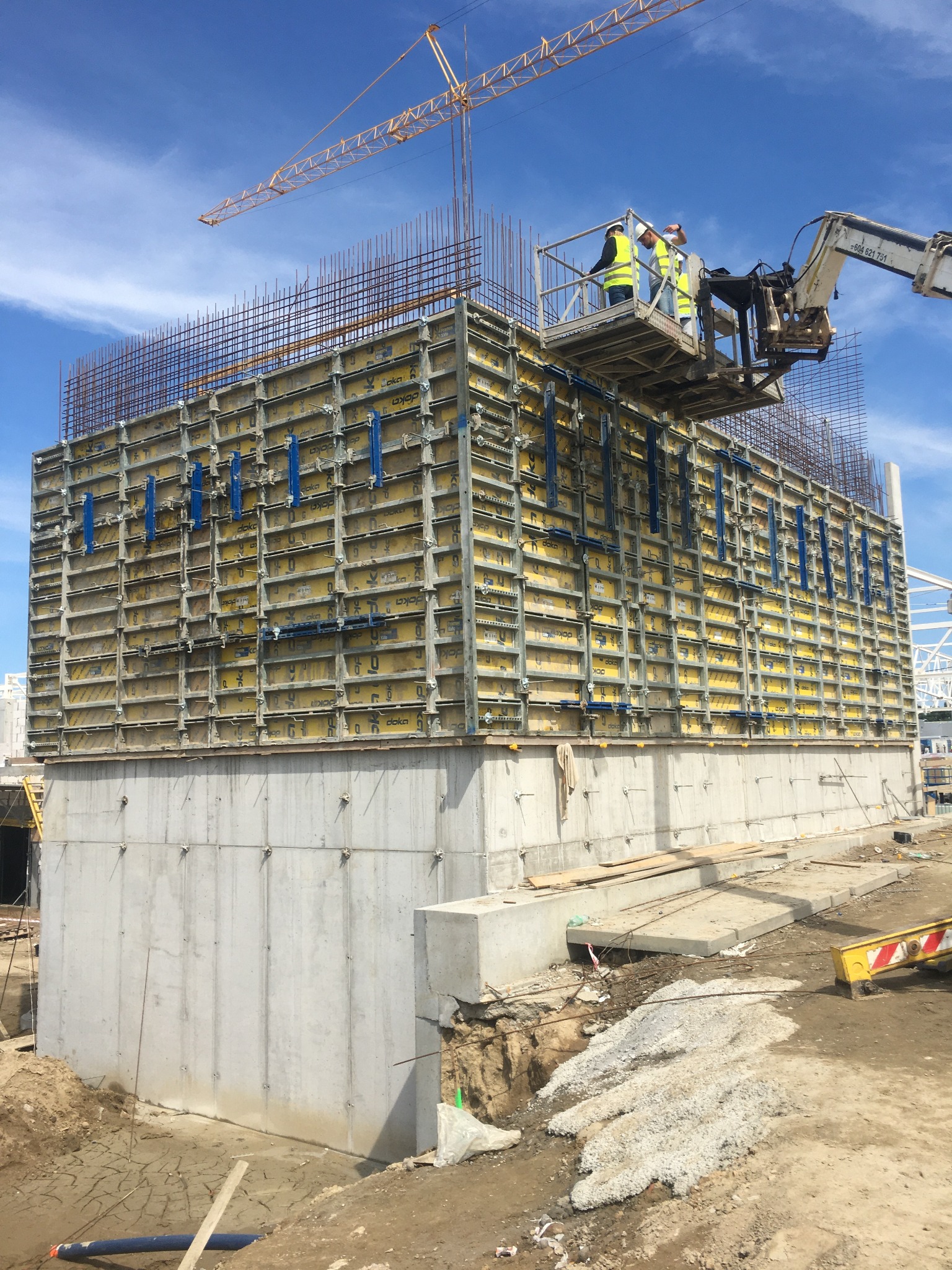 Wznoszenie ściany żelbetowej z szalunkiem systemowym Doka, widoczni dwaj pracownicy w podnośniku koszowym, zbrojenie pionowe wystające ponad szalunek, żuraw budowlany w tle.
