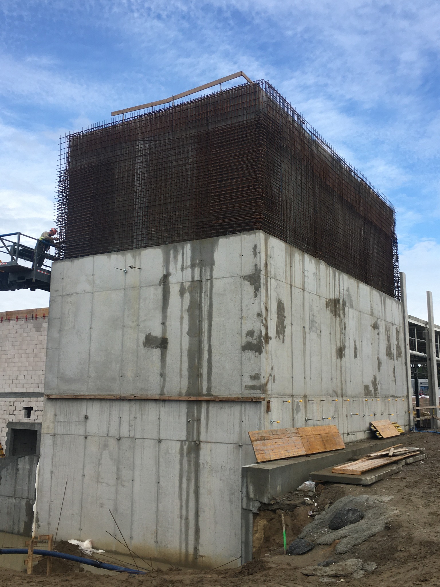 Wysoki budynek żelbetowy w trakcie budowy, z widocznym szkieletem zbrojeniowym na górnej części konstrukcji i pracownikiem na podnośniku koszowym w tle.