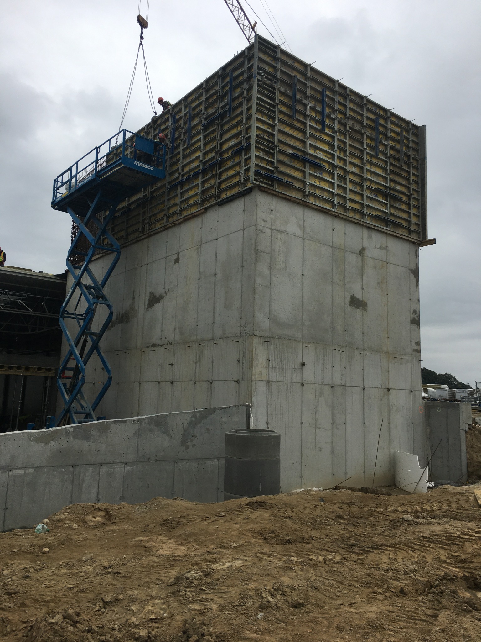 Wznoszenie żelbetowej konstrukcji: szalunek z żółtymi elementami, pracownicy na podnośniku koszowym i dźwig podnoszący elementy na tle pochmurnego nieba.