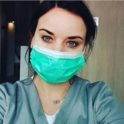 Portret kobiety w masce medycznej i szarym uniformie, z widocznym srebrnym naszyjnikiem z dwoma splecionymi kółkami.