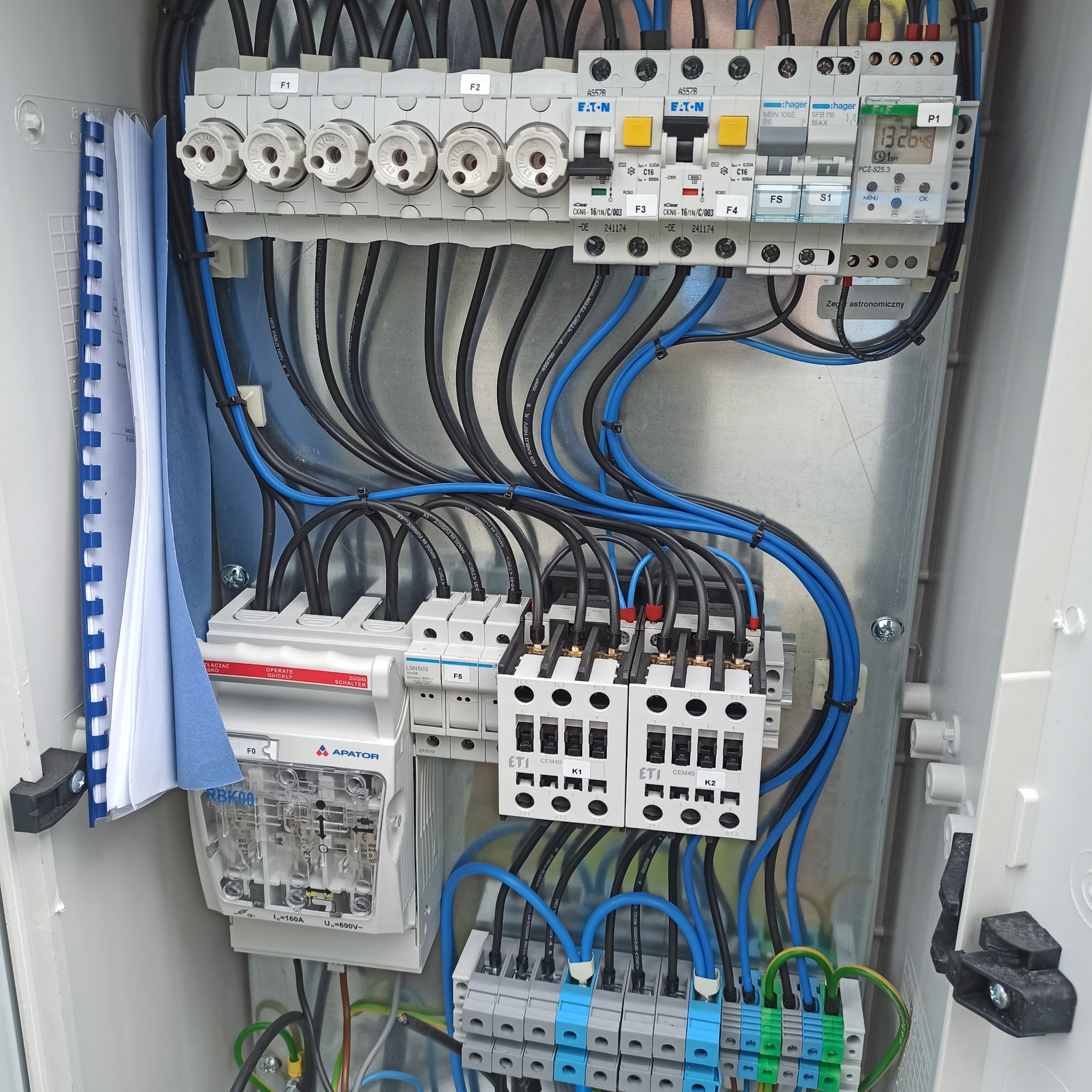 Otwarta skrzynka elektryczna z bezpiecznikami, wyłącznikami, przewodami czarnymi i niebieskimi oraz dokumentacją techniczną w segregatorze po lewej stronie. Widoczny zegar astronomiczny Hager...