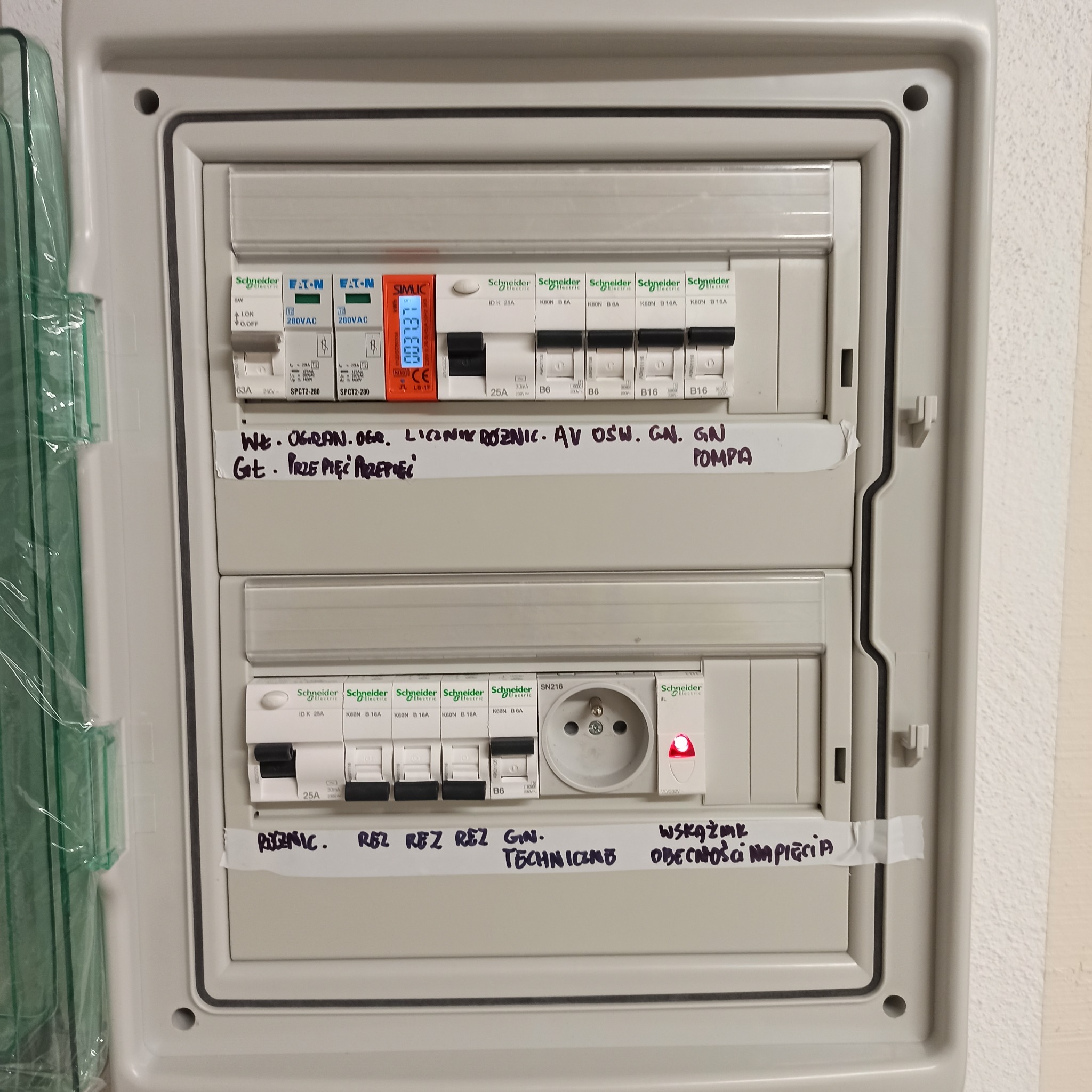 Otwarta skrzynka bezpiecznikowa z widocznymi bezpiecznikami, miernikiem cyfrowym, gniazdkiem i naklejonymi etykietami z opisami obwodów, widok z bliska.