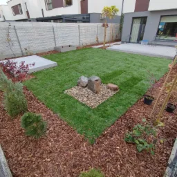 Widok z góry na nowocześnie urządzony ogród z równo przystrzyżonym trawnikiem, ozdobnymi kamieniami, korą drzewną, młodymi drzewkami i kostką brukową w tle zabudowy jednorodzinnej.