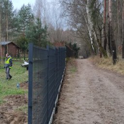 Usługi ogrodnicze Robert Starosz - Montaż panelowego ogrodzenia z metalowej siatki na działce z widocznym pracownikiem w odblaskowej kamizelce, narzędziami i ścieżką leśną w tle.