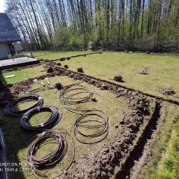 Usługi ogrodnicze Robert Starosz - Przygotowanie do instalacji podziemnego systemu nawadniania w ogrodzie: wykopane rowy, zwinięte rury, narzędzia ogrodnicze na trawniku, dom w tle.