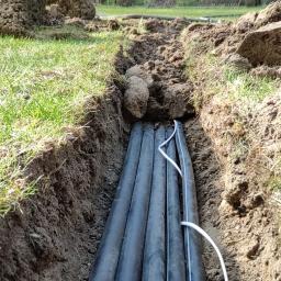 Usługi ogrodnicze Robert Starosz - Wykop z ułożonymi rurami do systemu nawadniania, widoczna biała instalacja elektryczna, ziemia i trawa na brzegach wykopu.