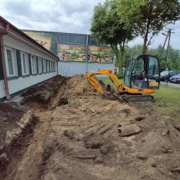 Usługi ogrodnicze Robert Starosz - Żółta mini koparka wykopuje wąski rów w ziemi wzdłuż budynku z białymi ścianami, przygotowanie terenu pod instalację.