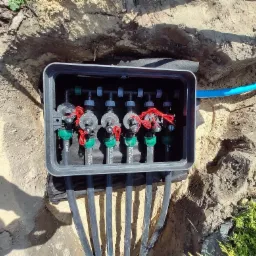 Skrzynka zaworowa z elektrozaworami zainstalowana w ziemi, widoczne czarne rury doprowadzające wodę do sekcji nawadniania, niebieski przewód zasilający, ziemia wokół skrzynki.