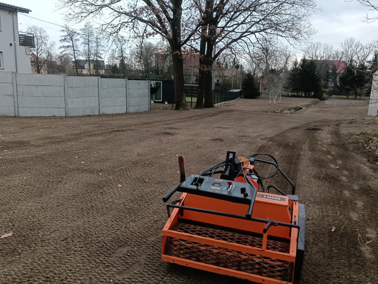 Maszyna do siewu trawy na przygotowanym terenie. Widoczne ślady po przejeździe, w tle domy i drzewa. Specyficzna faktura gleby po siewie.