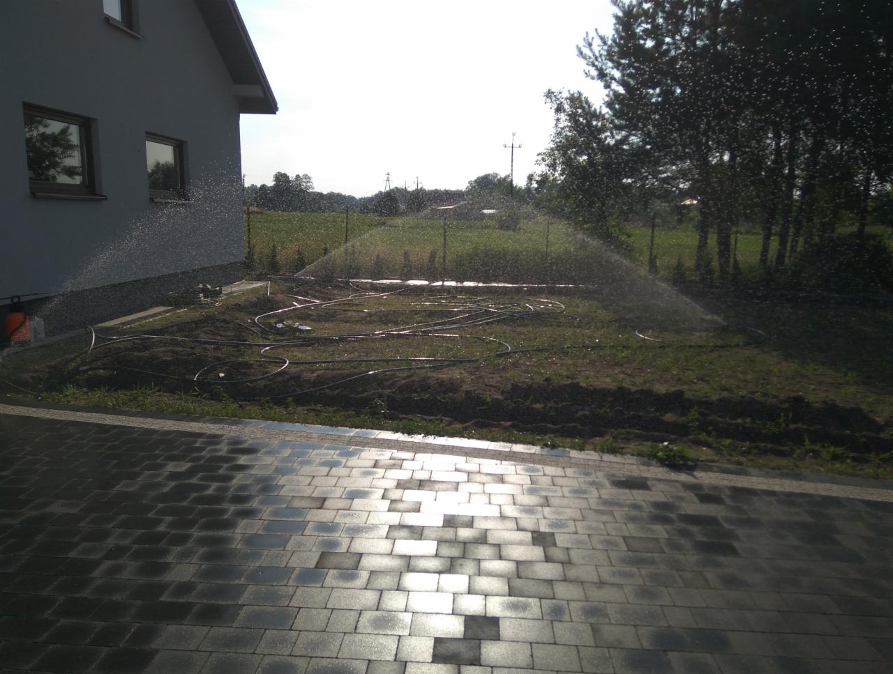 Instalacja zraszaczy ogrodowych w trakcie pracy, widoczne rury nawadniające na przygotowanym terenie, zraszacz rozpyla wodę w tle domu jednorodzinnego.