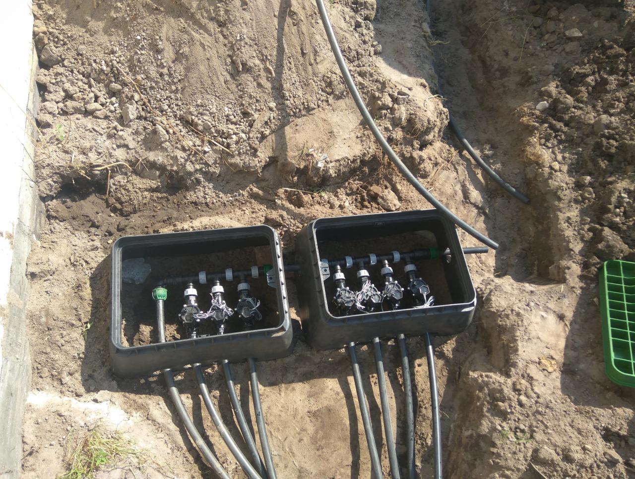 Dwa podziemne zawory nawadniające w czarnych skrzynkach, zainstalowane w wykopie ziemnym, widoczne rury doprowadzające wodę i zielony plastikowy pojemnik obok.