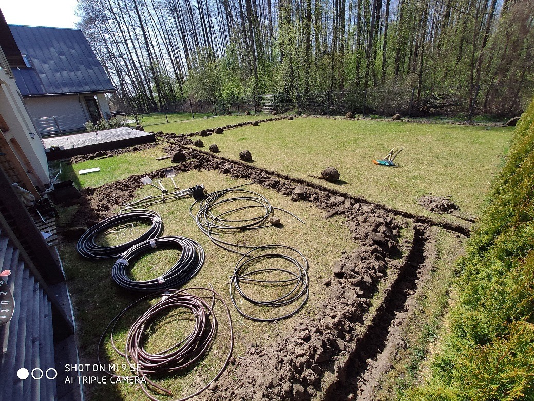 Przygotowanie do instalacji podziemnego systemu nawadniania w ogrodzie: wykopane rowy, zwinięte rury, narzędzia ogrodnicze na trawniku, dom w tle.
