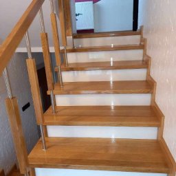 krzysztof gawor - Drewniane schody z jasnego drewna z białymi podstopnicami i metalowymi balustradami, widoczne z dołu, z fragmentem ściany w tle.