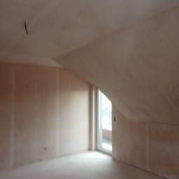 krzysztof gawor - Poddasze w stanie surowym, ściany pokryte jasną, jednolitą warstwą tynku gipsowego, widoczne łączenia płyt kartonowo-gipsowych, okno balkonowe wpuszczające naturalne światło.