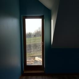 krzysztof gawor - Wnętrze poddasza z oknem w drewnianej ramie, ściany pomalowane na niebiesko, widoczny fragment drewnianej podłogi i schodów za oknem.