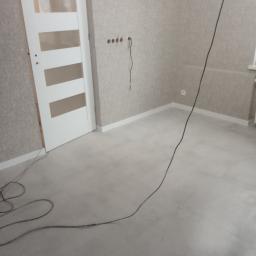 krzysztof gawor - Wnętrze pokoju po remoncie z zamontowaną białą, nowoczesną stolarką drzwiową z poziomymi przeszkleniami, widoczne szpachlowane ściany w odcieniach beżu, jasna podłoga i wystające przewody elektryczne.