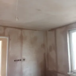 Surowe, jeszcze nie wyschnięte tynki na ścianach i suficie w nowo wykończonym pokoju z oknem i wystającymi kablami z sufitu.