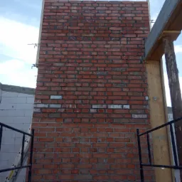 Wznoszona ściana z czerwonej cegły, widoczne fragmenty rusztowania i drewnianej konstrukcji szalunkowej, w tle niebieskie niebo i ściana z białych bloczków.