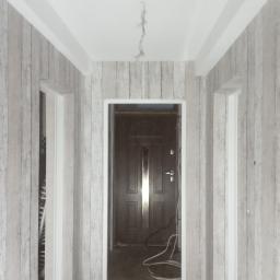 krzysztof gawor - Korytarz w trakcie remontu z tapetą imitującą drewno na ścianach, widoczne trzy otwory drzwiowe z białymi ościeżnicami, wystające kable z sufitu i przewody na podłodze.