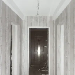 Korytarz w trakcie remontu z tapetą imitującą drewno na ścianach, widoczne trzy otwory drzwiowe z białymi ościeżnicami, wystające kable z sufitu i przewody na podłodze.