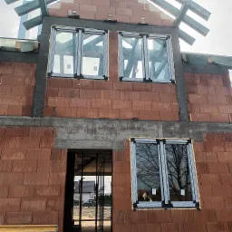 Surowy stan domu w trakcie budowy, widoczne ściany z czerwonej cegły, zamontowane okna z pianką montażową, betonowe nadproża i elementy konstrukcji dachu.