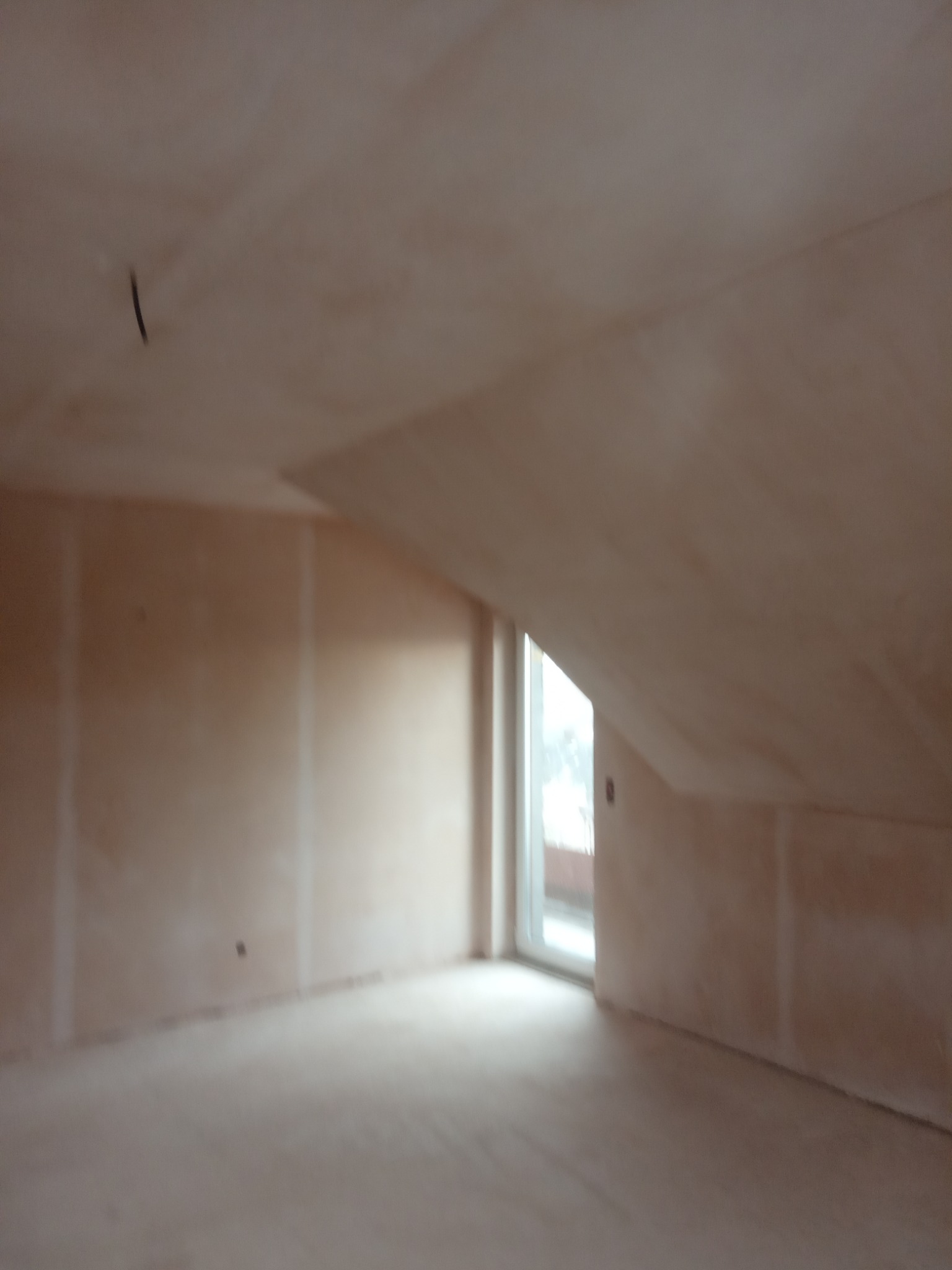 Poddasze w stanie surowym, ściany pokryte jasną, jednolitą warstwą tynku gipsowego, widoczne łączenia płyt kartonowo-gipsowych, okno balkonowe wpuszczające naturalne światło.