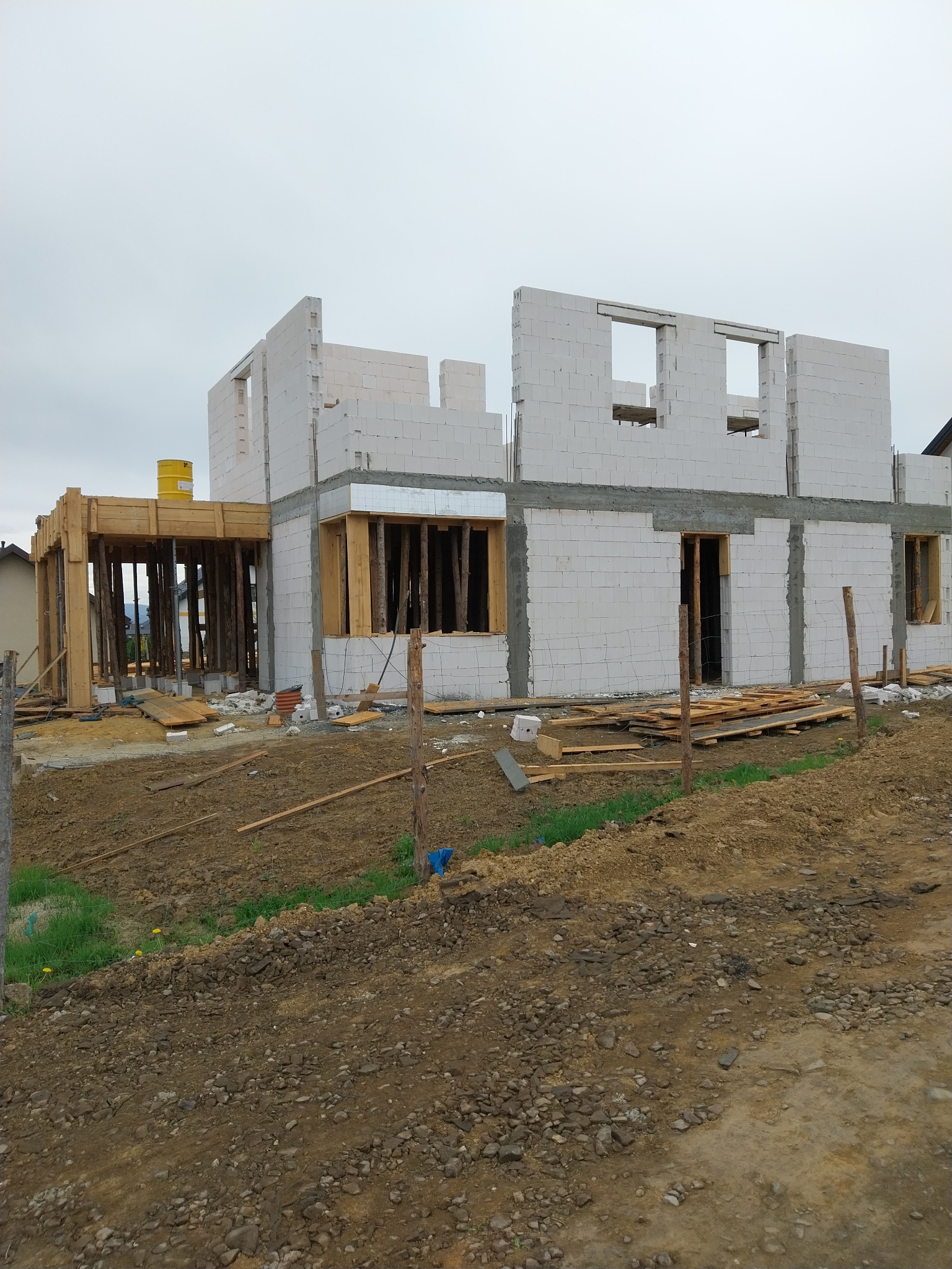 Budowa domu w trakcie: wzniesione ściany z bloczków, widoczne otwory okienne i drzwiowe, drewniane podpory. Teren wokół w stanie surowym.