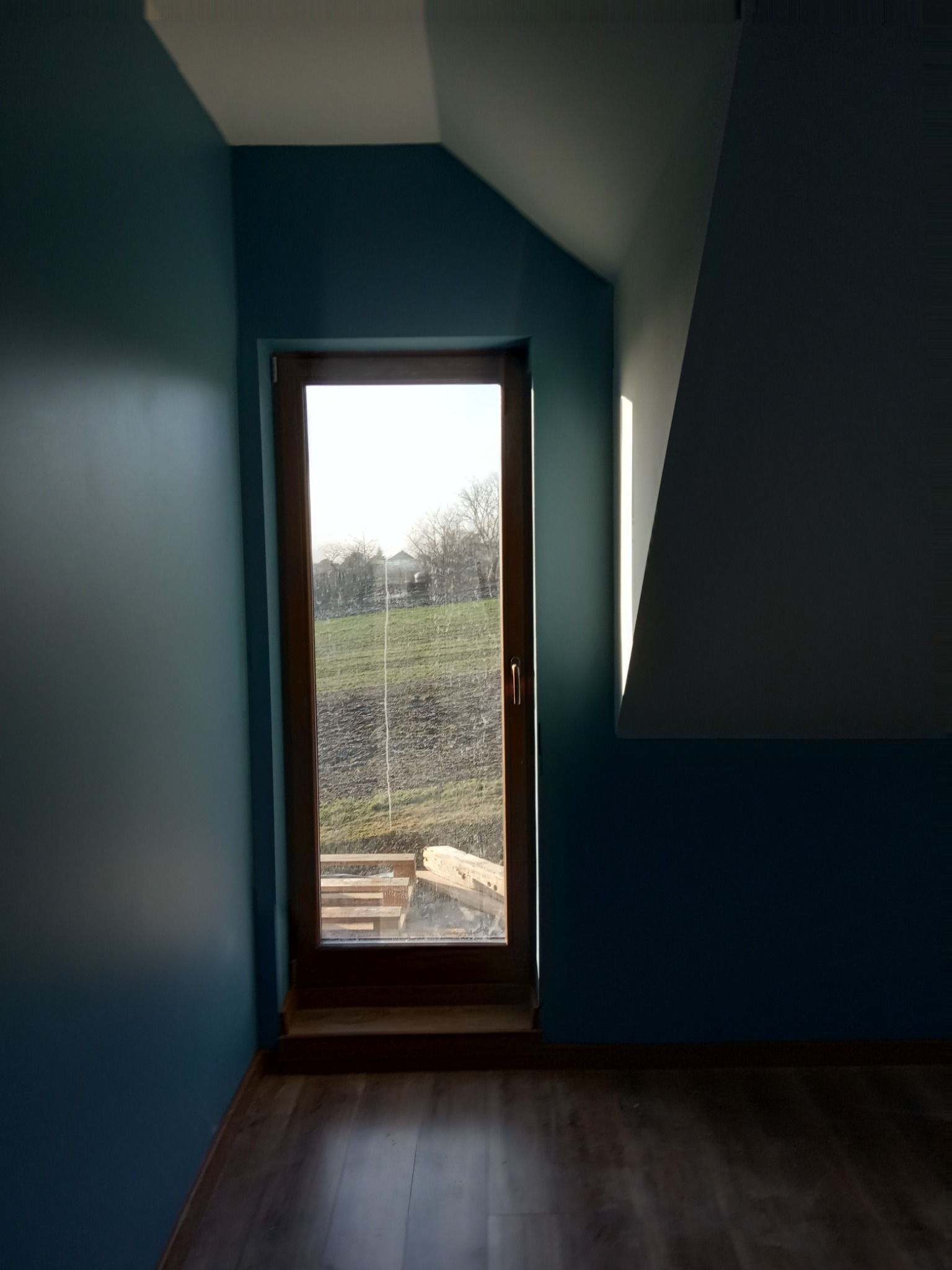 Wnętrze poddasza z oknem w drewnianej ramie, ściany pomalowane na niebiesko, widoczny fragment drewnianej podłogi i schodów za oknem.