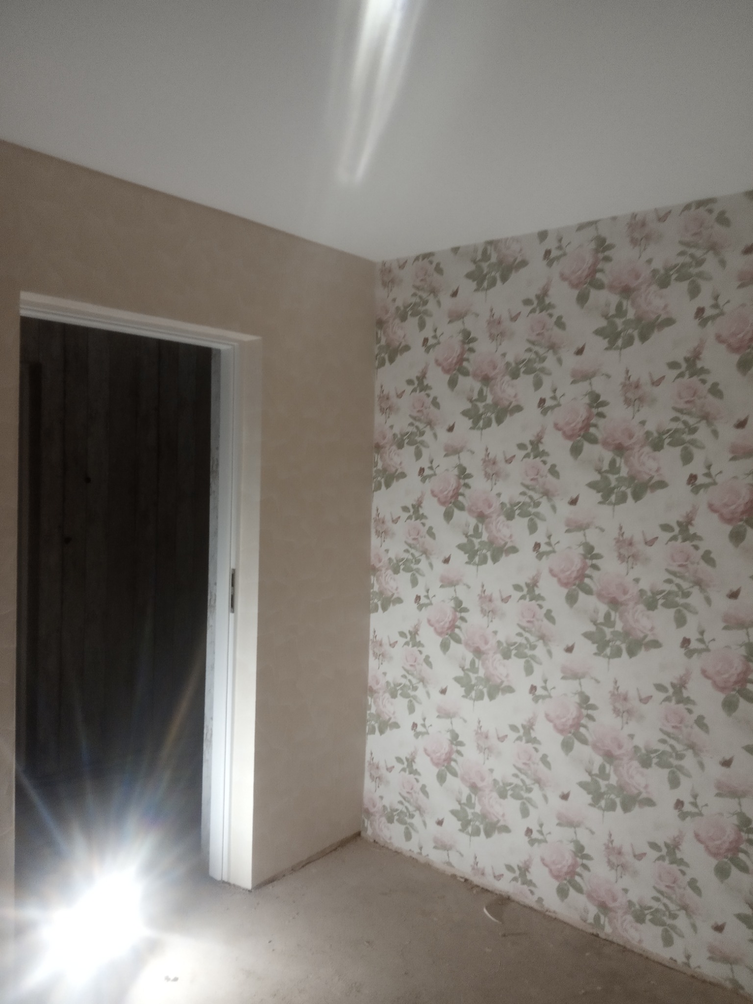 Pokój po remoncie: ściana z tapetą w różowe róże, druga ściana pokryta jasnym tynkiem strukturalnym, widoczne ościeżnice drzwiowe.