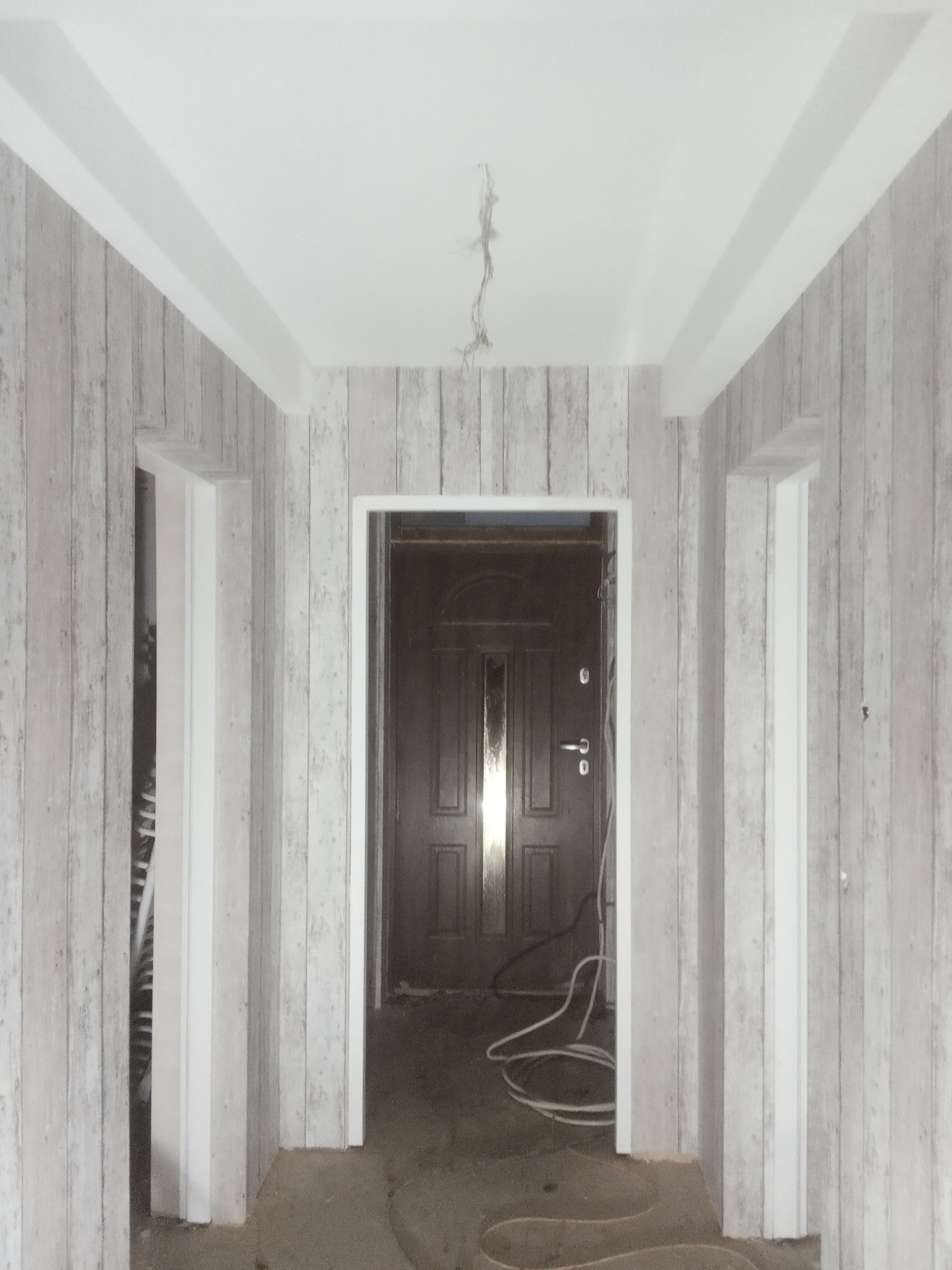 Korytarz w trakcie remontu z tapetą imitującą drewno na ścianach, widoczne trzy otwory drzwiowe z białymi ościeżnicami, wystające kable z sufitu i przewody na podłodze.