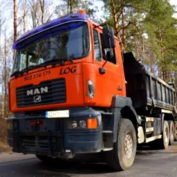 Pomarańczowa ciężarówka MAN z czarną skrzynią ładunkową, zaparkowana na asfaltowej drodze w otoczeniu drzew iglastych, z numerem telefonu i logo firmy na kabinie.