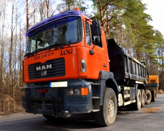 Pomarańczowa ciężarówka MAN z czarną skrzynią ładunkową, zaparkowana na asfaltowej drodze w otoczeniu drzew iglastych, z numerem telefonu i logo firmy na kabinie.