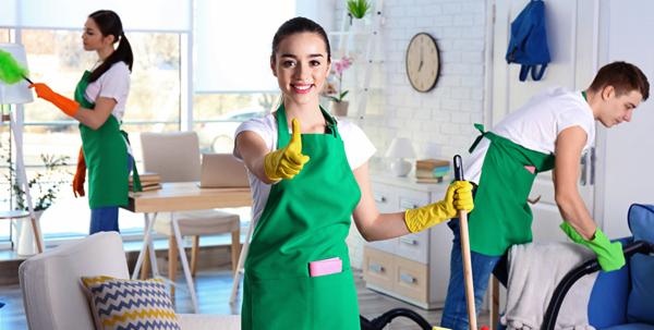 Zespół sprzątający w zielonych fartuchach: kobieta z uśmiechem i kciukiem w górę, mężczyzna odkurza sofę, a kobieta czyści półkę z kurzu w jasnym, zadbanym wnętrzu.