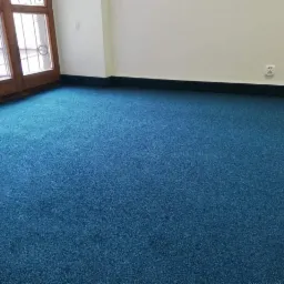 Pok&oacute;j z nową, intensywnie niebieską wykładziną dywanową, widoczne listwy przypodłogowe i drewniane okno z ozdobnymi szprosami.