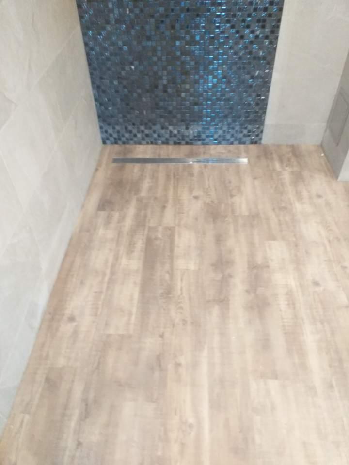 Jasna podłoga z paneli imitujących drewno w łazience, z odpływem liniowym i mozaiką na ścianie.