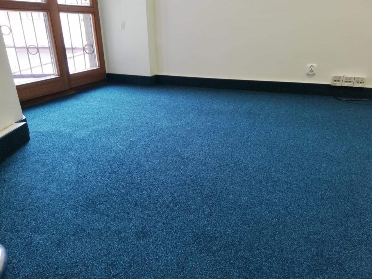 Pokój z nową, intensywnie niebieską wykładziną dywanową, widoczne listwy przypodłogowe i drewniane okno z ozdobnymi szprosami.