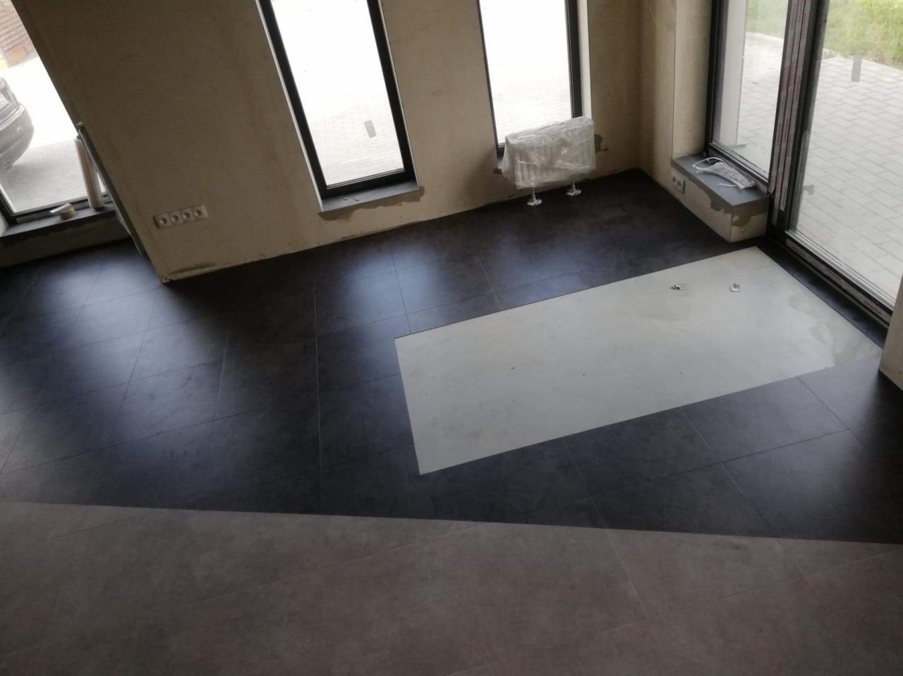 Wnętrze w trakcie prac wykończeniowych; położona ciemna, kwadratowa podłoga z płytek, fragment białej płyty w podłodze, surowe ściany i okna w czarnych ramach.